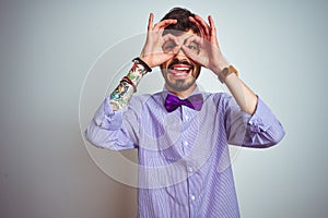 Young man with tattoo wearing purple shirt and bow tie over isolated white background doing ok gesture like binoculars sticking