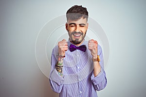 Young man with tattoo wearing purple shirt and bow tie over isolated white background excited for success with arms raised and