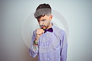 Young man with tattoo wearing purple shirt and bow tie over isolated white background feeling unwell and coughing as symptom for