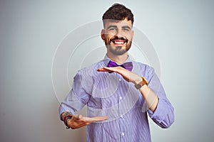 Young man with tattoo wearing purple shirt and bow tie over isolated white background gesturing with hands showing big and large