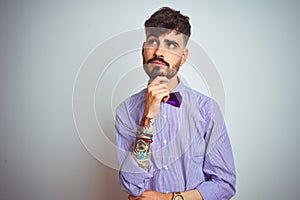 Young man with tattoo wearing purple shirt and bow tie over isolated white background with hand on chin thinking about question,