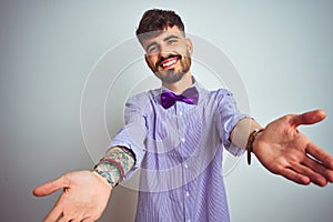 Young man with tattoo wearing purple shirt and bow tie over isolated white background looking at the camera smiling with open arms