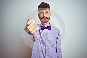 Young man with tattoo wearing purple shirt and bow tie over isolated white background looking unhappy and angry showing rejection