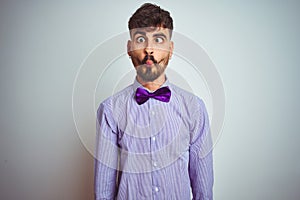 Young man with tattoo wearing purple shirt and bow tie over isolated white background making fish face with lips, crazy and