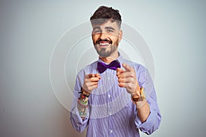 Young man with tattoo wearing purple shirt and bow tie over isolated white background pointing fingers to camera with happy and