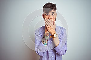 Young man with tattoo wearing purple shirt and bow tie over isolated white background shocked covering mouth with hands for