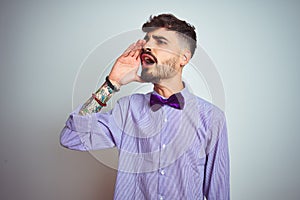 Young man with tattoo wearing purple shirt and bow tie over isolated white background shouting and screaming loud to side with