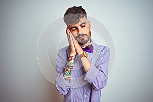 Young man with tattoo wearing purple shirt and bow tie over isolated white background sleeping tired dreaming and posing with
