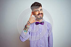 Young man with tattoo wearing purple shirt and bow tie over isolated white background smiling doing phone gesture with hand and
