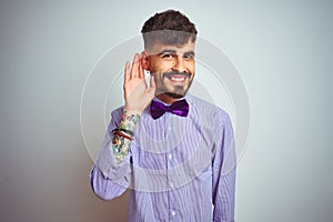 Young man with tattoo wearing purple shirt and bow tie over isolated white background smiling with hand over ear listening an