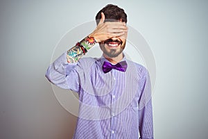 Young man with tattoo wearing purple shirt and bow tie over isolated white background smiling and laughing with hand on face