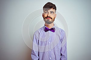 Young man with tattoo wearing purple shirt and bow tie over isolated white background smiling looking to the side and staring away