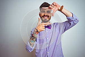 Young man with tattoo wearing purple shirt and bow tie over isolated white background smiling making frame with hands and fingers