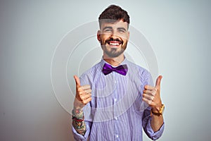 Young man with tattoo wearing purple shirt and bow tie over isolated white background success sign doing positive gesture with