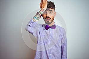Young man with tattoo wearing purple shirt and bow tie over isolated white background surprised with hand on head for mistake,
