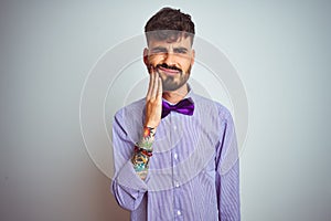 Young man with tattoo wearing purple shirt and bow tie over isolated white background touching mouth with hand with painful