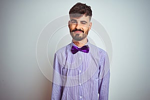 Young man with tattoo wearing purple shirt and bow tie over isolated white background winking looking at the camera with sexy