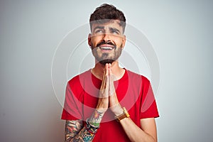 Young man with tattoo wearing red t-shirt standing over isolated white background begging and praying with hands together with