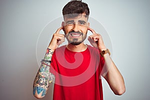 Young man with tattoo wearing red t-shirt standing over isolated white background covering ears with fingers with annoyed