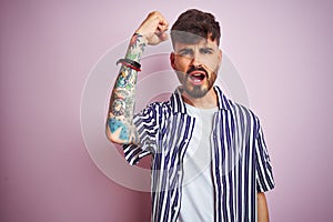 Young man with tattoo wearing striped shirt standing over isolated pink background angry and mad raising fist frustrated and