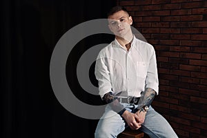 Young man with tattoos sitting on stool indoors. Space for text