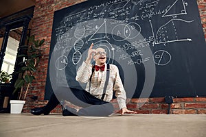 Young man, teacher, scientist sitting by blackboard with scientific calculations and formulas, finding out solution