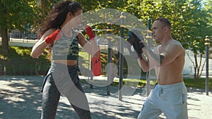 Young man teaches beautiful woman boxing blows outdoors at sunny day