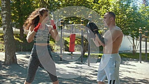 Young man teaches beautiful woman boxing blows outdoors at sunny day, slow-motion