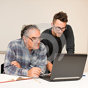 Young man teaching eldery man of usage of computer. Intergenerational transfer of computer skills.