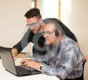 Young man teaching eldery man of usage of computer. Intergenerational transfer of computer skills.