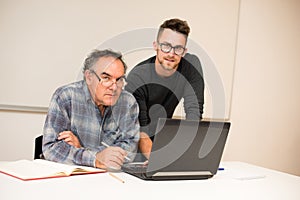 Young man teaching eldery man of usage of computer. Intergenerational transfer of computer skills.