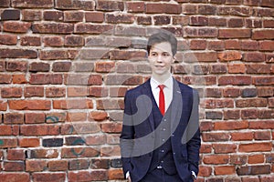 A young man, a teenager, in a classic suit. Pondering is standing in front of the old wall of red brick, putting his hands in his