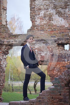 A young man, a teenager, in a classic suit. Pondering is standing in front of the old wall of red brick, putting his hands in his