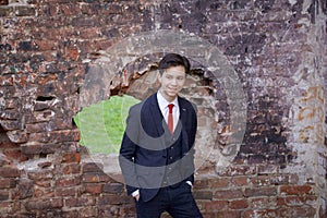 A young man, a teenager, in a classic suit. Pondering is standing in front of the old wall of red brick, putting his hands in his