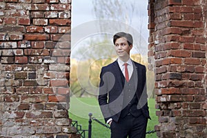 A young man, a teenager, in a classic suit. Pondering is standing in front of the old wall of red brick, putting his hands in his