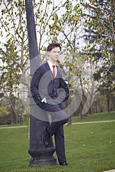 A young man, a teenager, in a classic suit. Relying on a vintage lantern in the park. Put your hands in your pockets.