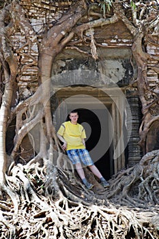Young man, the teenager at an entrance to the destroyed covered with roots of trees temple Prasat Chrap in the Koh Ker temple comp