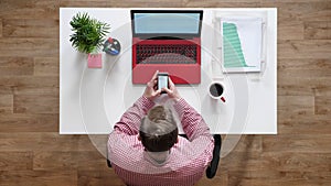 Young man texting on phone, topshot, sitting behind desk with laptop and coffee