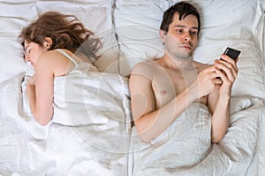 Young man is texting with someone using phone while his wife is sleeping near him.