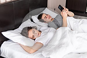 Young man is texting with someone using phone while his wife is sleeping near him. Chatting concept