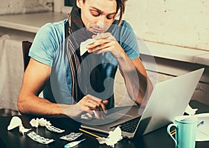 Young man with thermometer sick blowing his nose in his living room working notebook