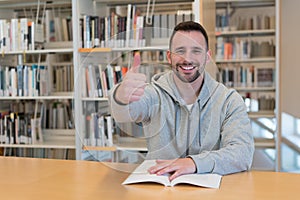 Young man with thumbs up happy and smiling with a book on the table in the library