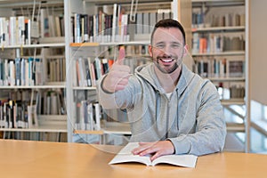 Joven hombre pulgar arriba feliz a un libro sobre el mesa en biblioteca 