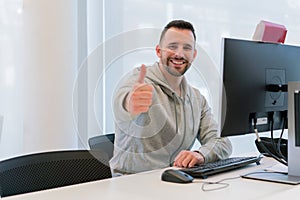 Young man with thumbs up happy and smiling for having achieved his goals in the office in front of the computer