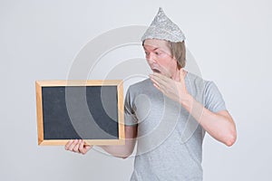 Young man with tin foil hat holding blackboard and looking shocked