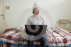 Young man with tin foil hat thinking while using laptop in the bedroom