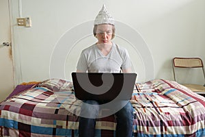 Young man with tin foil hat using laptop in the bedroom