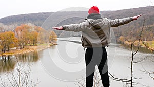 A young man at the top of the mountain imitates an airplane. Happy man in nature by the river with outstretched arms.