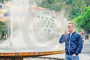 Young man tourist with blue jacket look at camera and drink therapeutic mineral water from cup