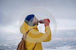 Young man tourist hiker standing on mountain top drinking hot tea from thermos during winter hiking